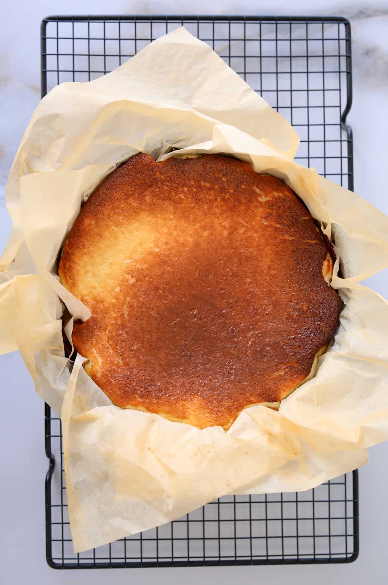 A golden-brown, slightly burnt cheesecake sits on parchment paper in a round baking pan, cooling on a wire rack.