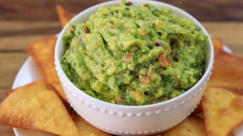A white bowl filled with chunky guacamole sits on a plate surrounded by crispy tortilla chips, all placed on a wooden surface.