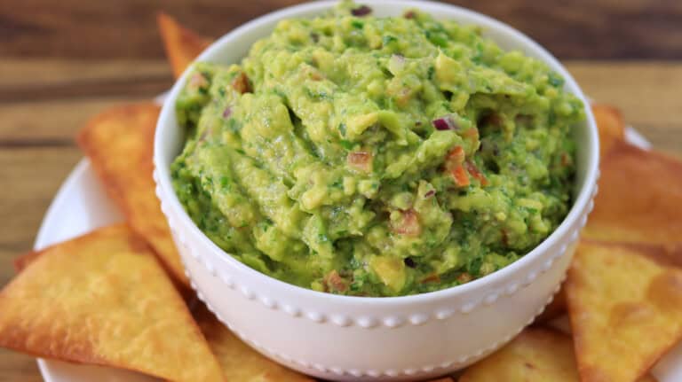 A white bowl filled with chunky guacamole sits on a plate surrounded by crispy tortilla chips, all placed on a wooden surface.