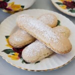 Three ladyfinger biscuits dusted with powdered sugar are arranged on a decorative plate with yellow and red floral designs. Other similar plates are partially visible in the background.