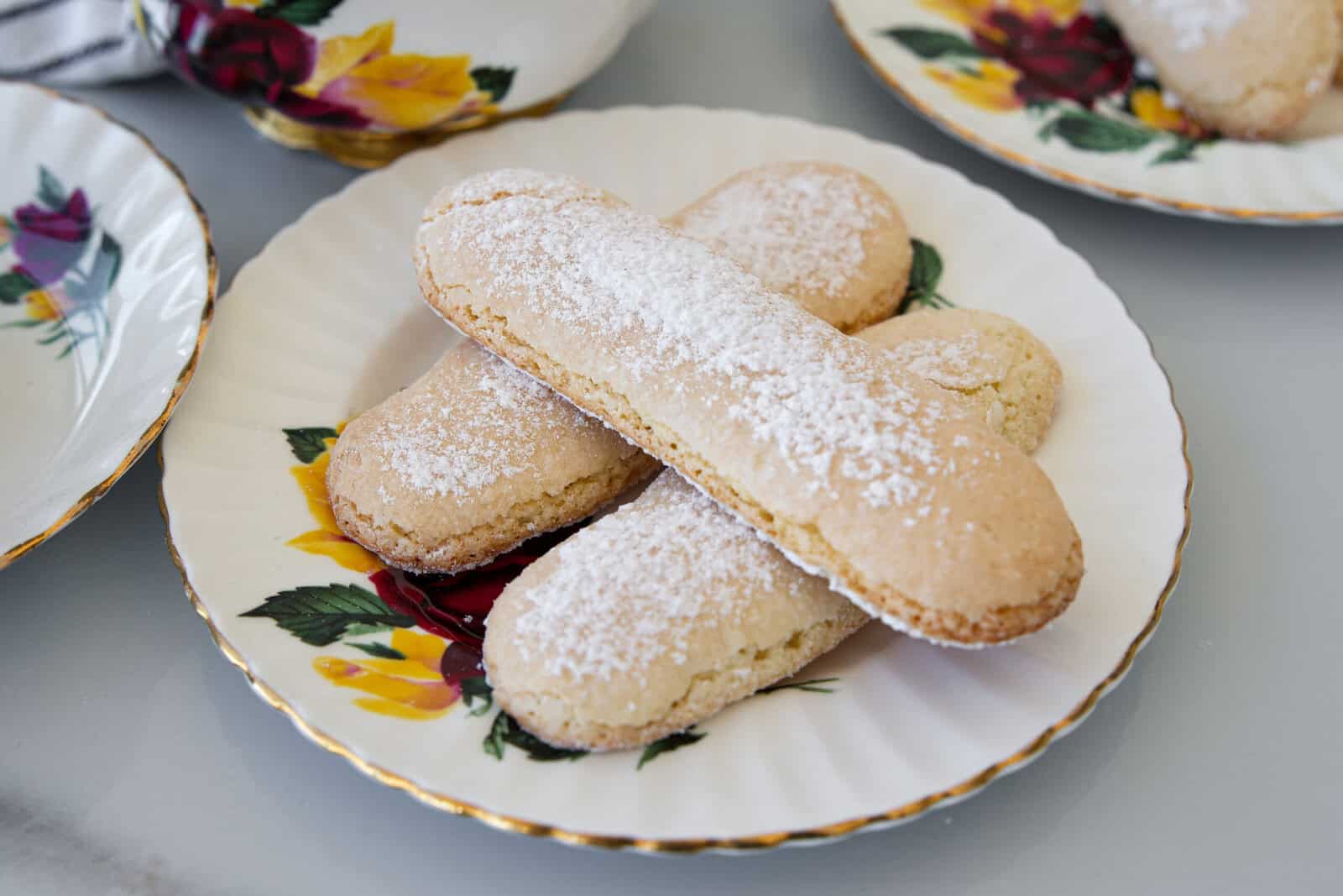 Three ladyfinger biscuits dusted with powdered sugar are arranged on a decorative plate with yellow and red floral designs. Other similar plates are partially visible in the background.