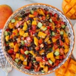 A colorful bowl of mango black bean salad with diced mango, black beans, cucumber, red bell pepper, tomato, and red onion, surrounded by fresh mangoes and salad utensils on a marble surface.