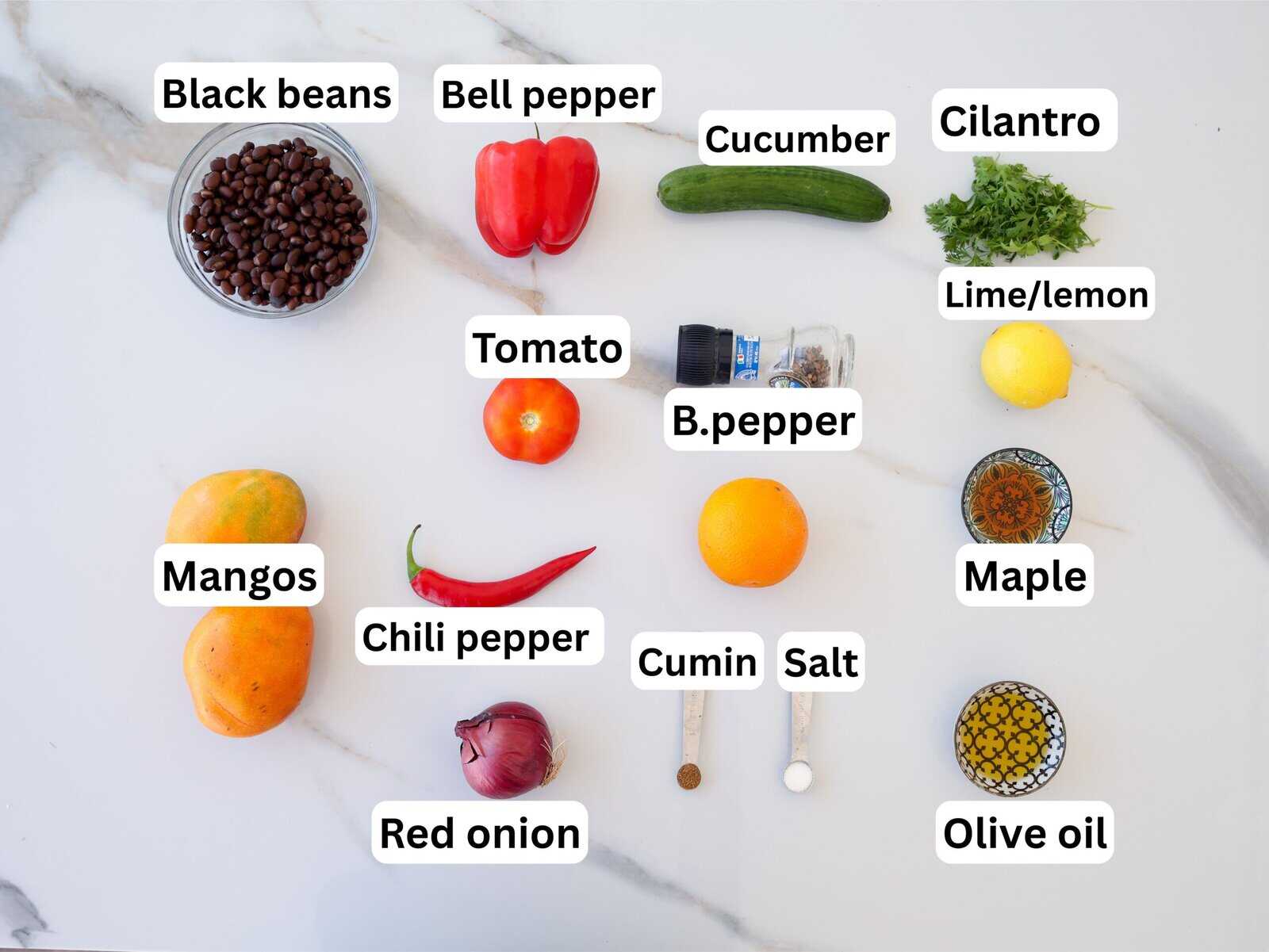 A flat lay of ingredients on a white surface, each labeled: black beans, bell pepper, cucumber, cilantro, lime/lemon, tomato, black pepper, mangoes, chili pepper, orange, cumin, salt, maple syrup, olive oil, and red onion.