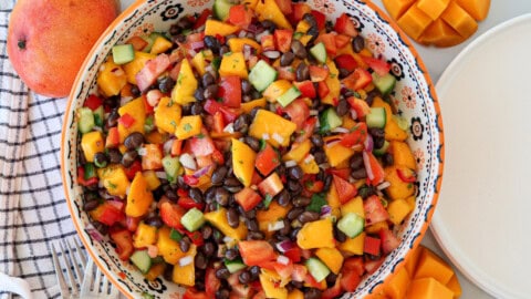 Easy Black Bean Mango Salad with Lime Dressing A colorful salad with diced mango, black beans, cucumber, red pepper, tomato, and red onion in a decorative bowl, surrounded by fresh mango and a striped napkin.