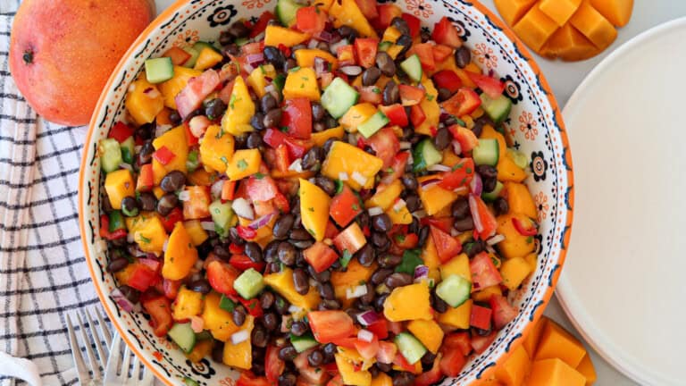 A colorful salad with diced mango, black beans, cucumber, red pepper, tomato, and red onion in a decorative bowl, surrounded by fresh mango and a striped napkin.