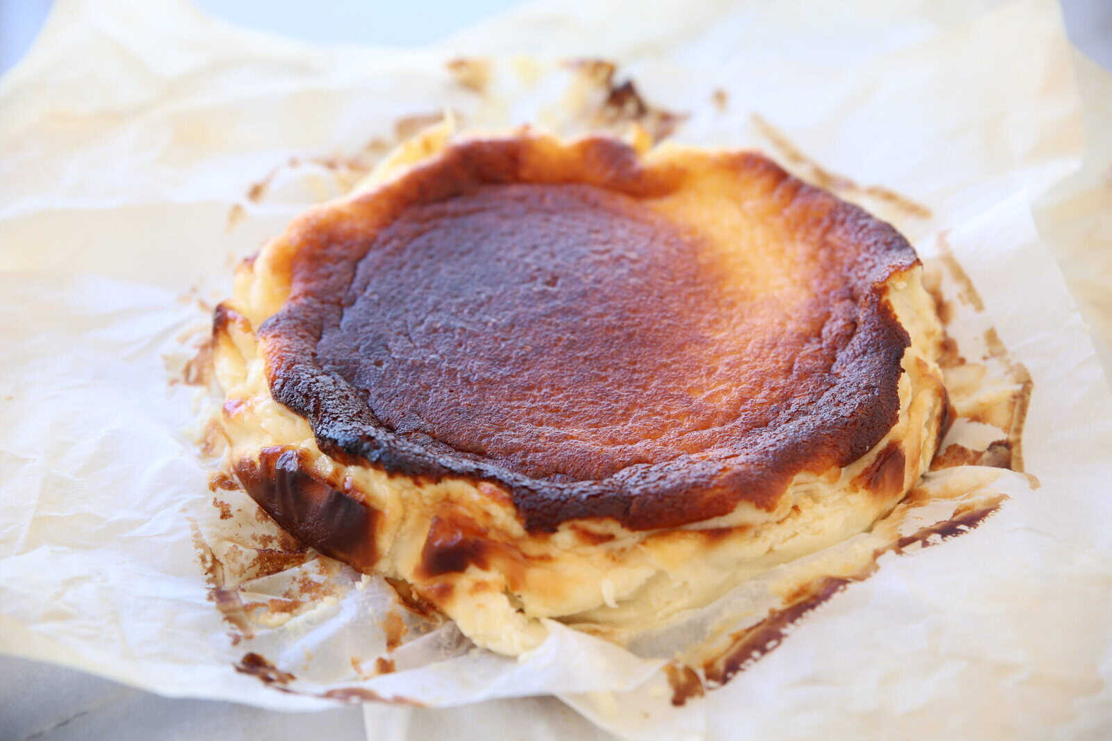 A rustic Basque burnt cheesecake with a caramelized top and creamy edges sits on crumpled parchment paper. The outer layer is browned and cracked, contrasting with the lighter, creamy inside.