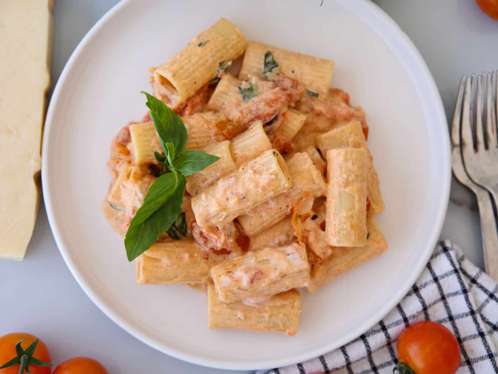A white plate of rigatoni pasta in a creamy tomato sauce, garnished with fresh basil. The plate is on a table with a checkered cloth, cherry tomatoes, cutlery, and a wedge of cheese nearby.