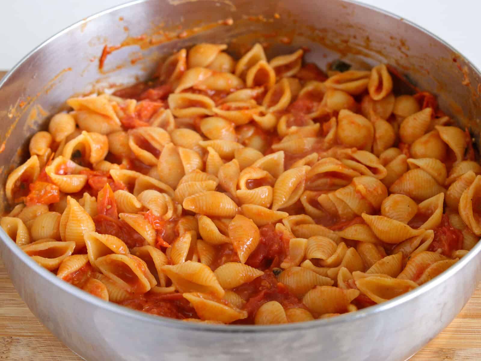 A close-up of a metal pot filled with shell pasta mixed in a rich cherry tomato sauce, sitting on a wooden surface.