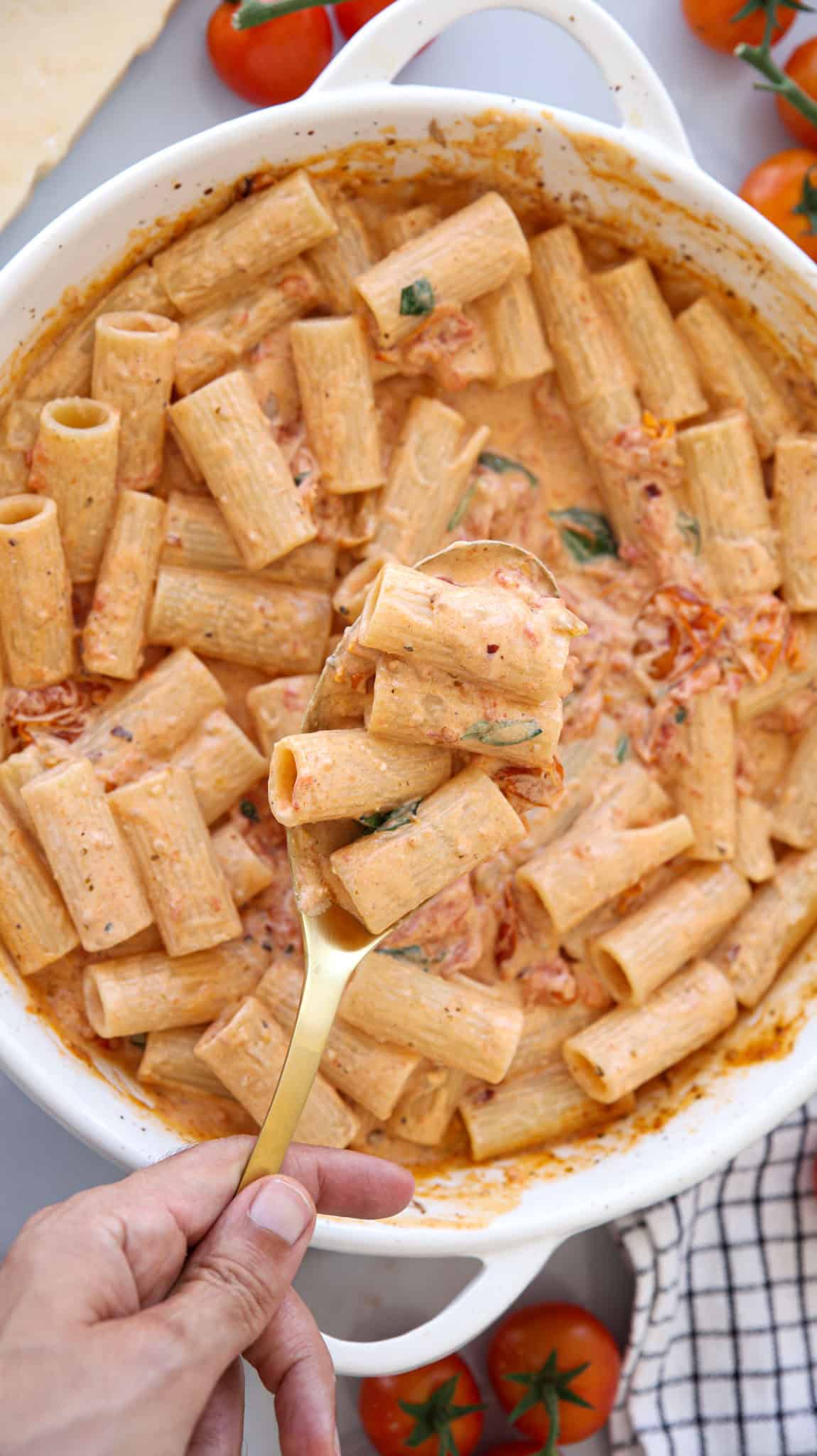 A hand holding a gold spoon scoops creamy rigatoni pasta with tomato mascarpone sauce and herbs from a white dish, surrounded by cherry tomatoes.