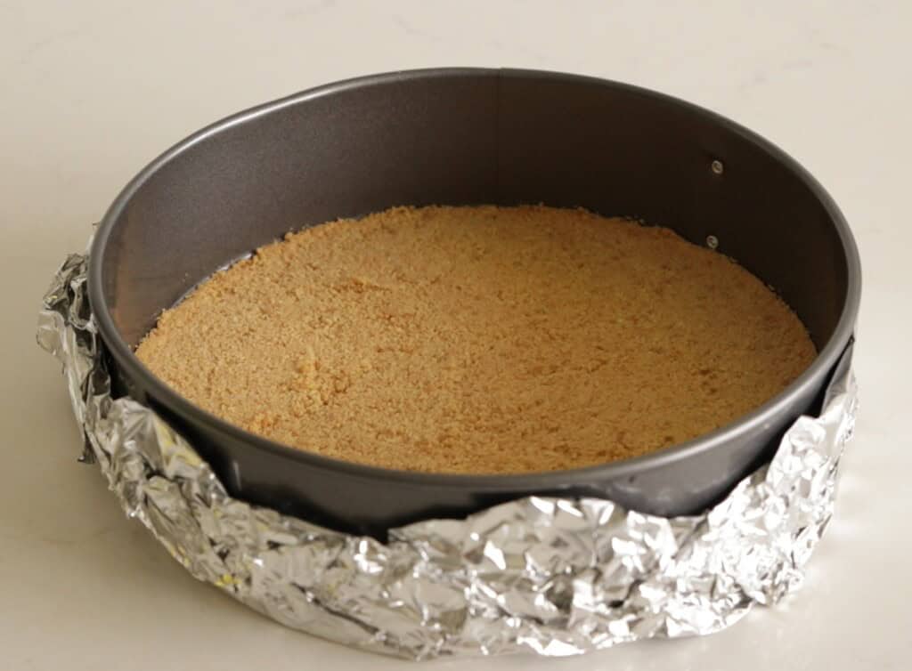 A round baking pan with foil wrapped around the outside contains a pressed layer of crumbly graham cracker crust, ready for baking or filling.