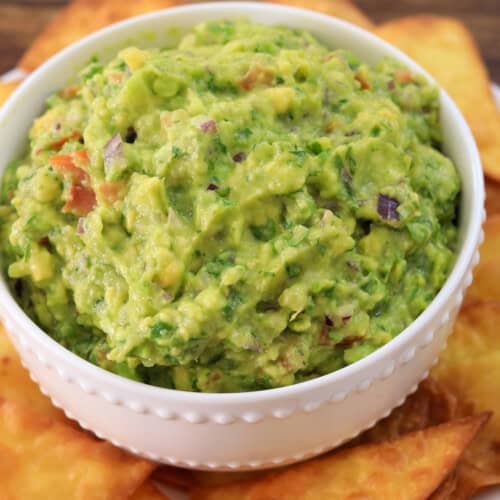 A white bowl filled with chunky guacamole, garnished with herbs, sits on a plate surrounded by golden, crispy tortilla chips on a wooden table.