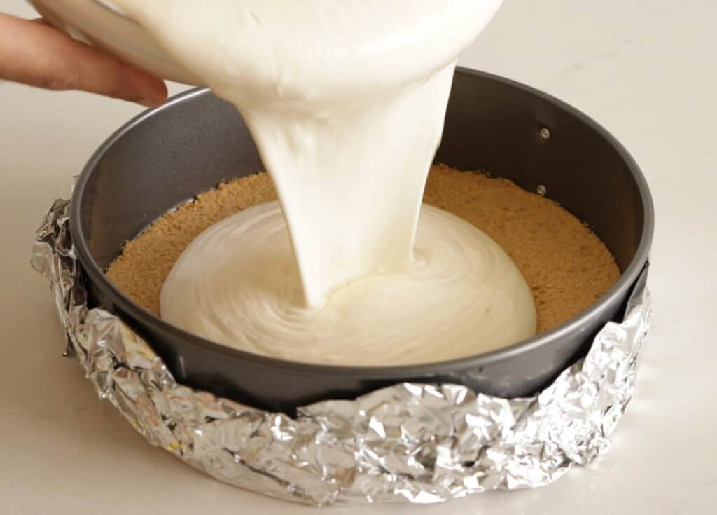 A close-up of cheesecake batter being poured into a round, foil-wrapped springform pan with a graham cracker crust on a light surface.