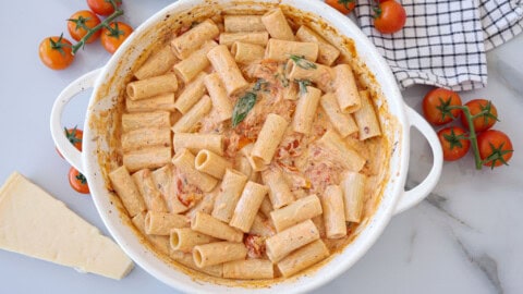 A white dish filled with rigatoni pasta in a creamy cherry tomato sauce with mascarpone, garnished with basil. Fresh tomatoes, a block of cheese, and a checkered cloth are nearby on a marble surface.