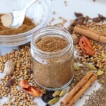 A glass jar filled with ground spice mix sits on a white surface surrounded by whole spices, including cinnamon sticks, cardamom pods, coriander seeds, black pepper, nutmeg, and star anise. A bowl of mixed spices is in the background.
