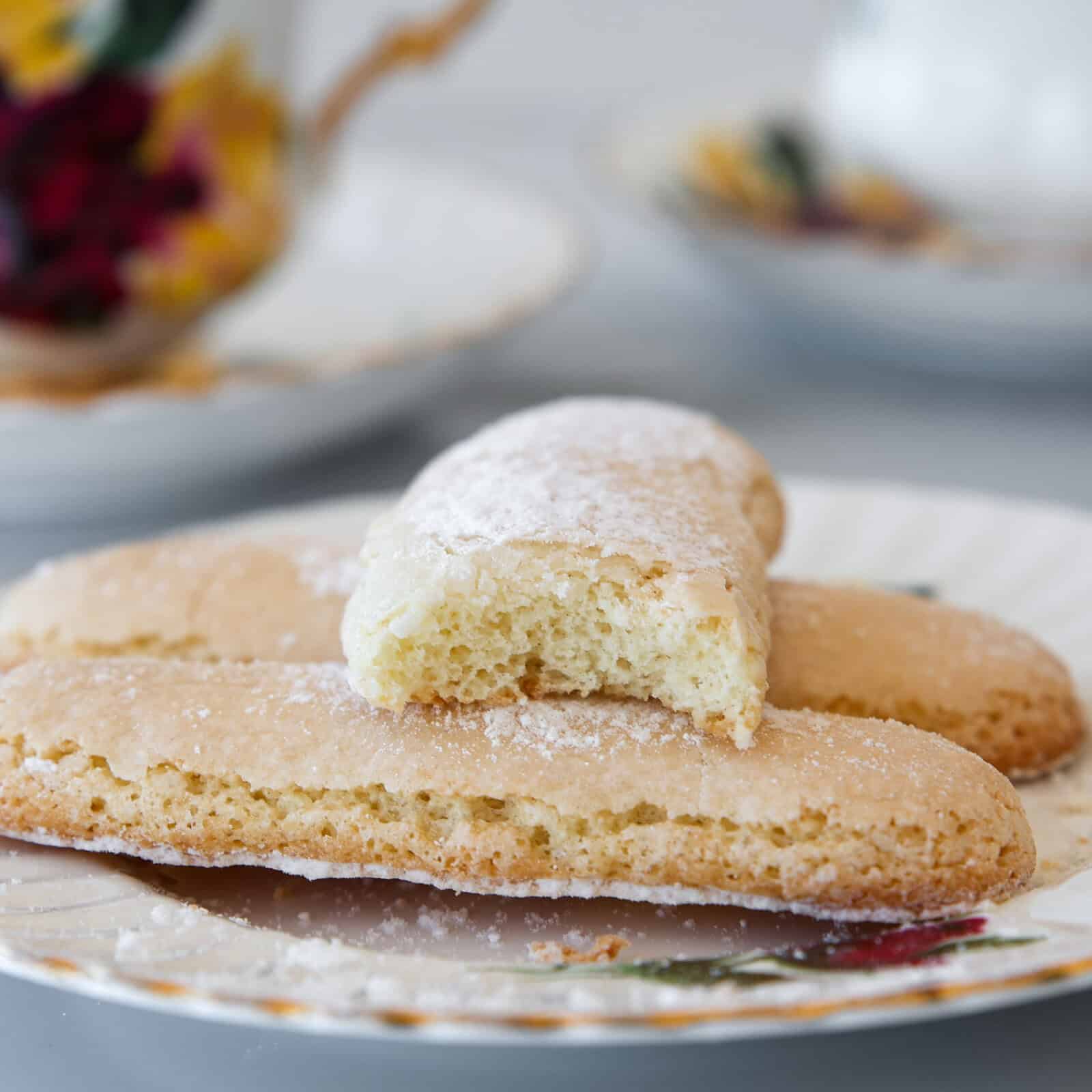 Three ladyfinger biscuits dusted with powdered sugar are arranged on a decorative plate, with one biscuit placed on top and partially bitten to reveal its airy interior. A blurred teacup and saucer are in the background.