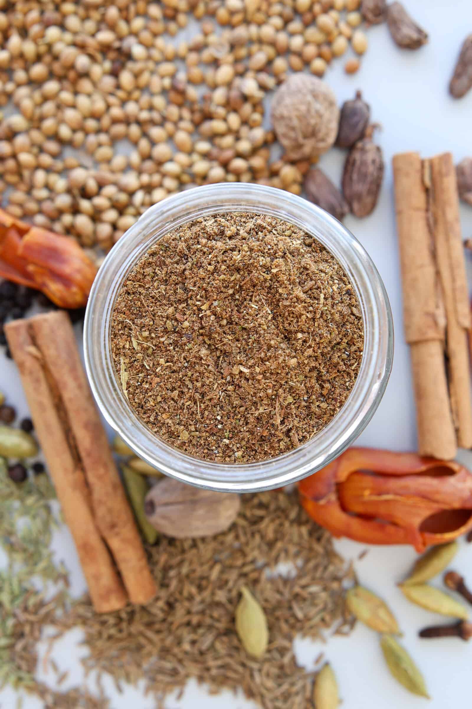 A jar filled with garam masala spice blend sits surrounded by whole spices, including cinnamon sticks, coriander seeds, nutmeg, cardamom pods, mace, cumin seeds, and black peppercorns on a white surface.