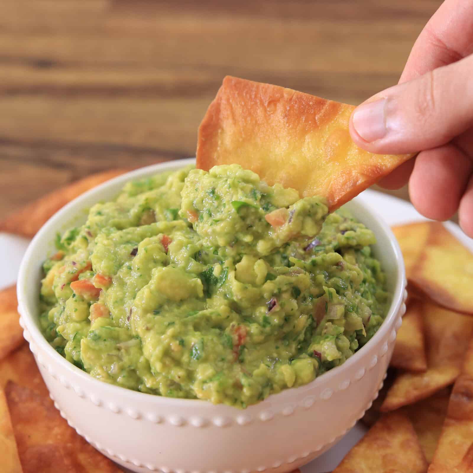 A hand dips a crispy chip into a bowl of chunky guacamole, surrounded by more chips on a plate, set on a wooden table.