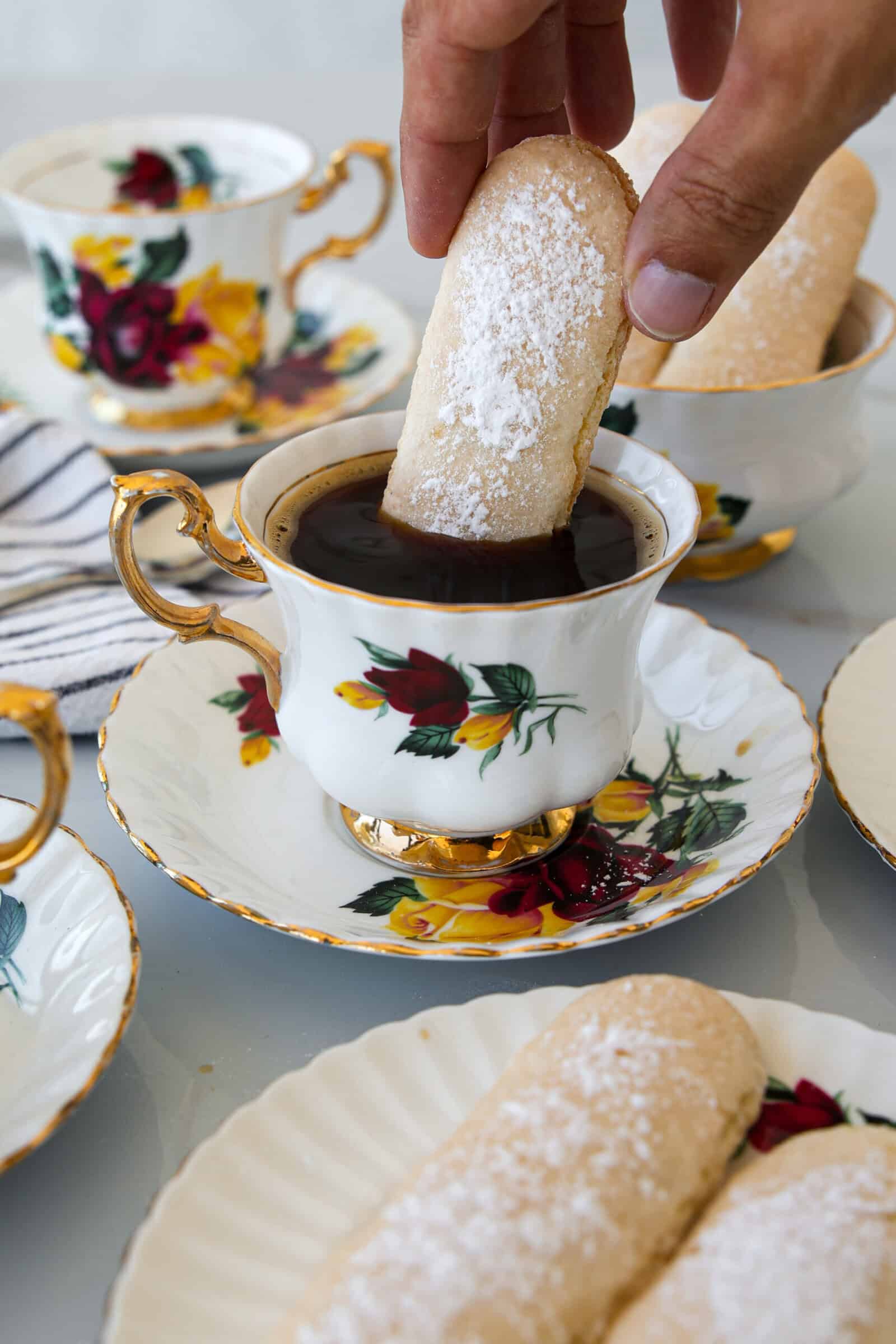 A hand dips a powdered sugar-covered ladyfinger cookie into a cup of black coffee, served in a floral-patterned teacup with matching saucers and plates, which also hold more ladyfinger cookies.