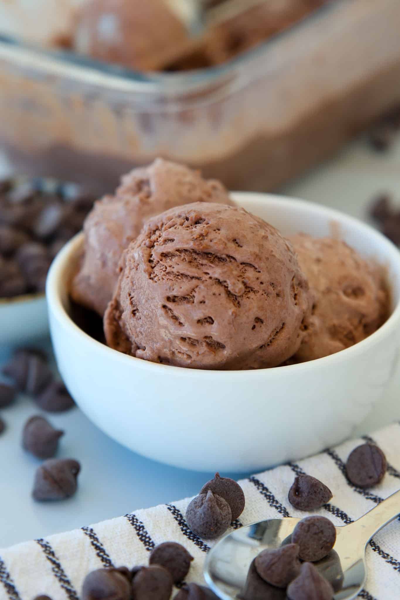 A white bowl filled with scoops of 3-ingredient chocolate ice cream sits on a striped cloth, surrounded by scattered chocolate chips, with a spoon and a container of more ice cream in the background.