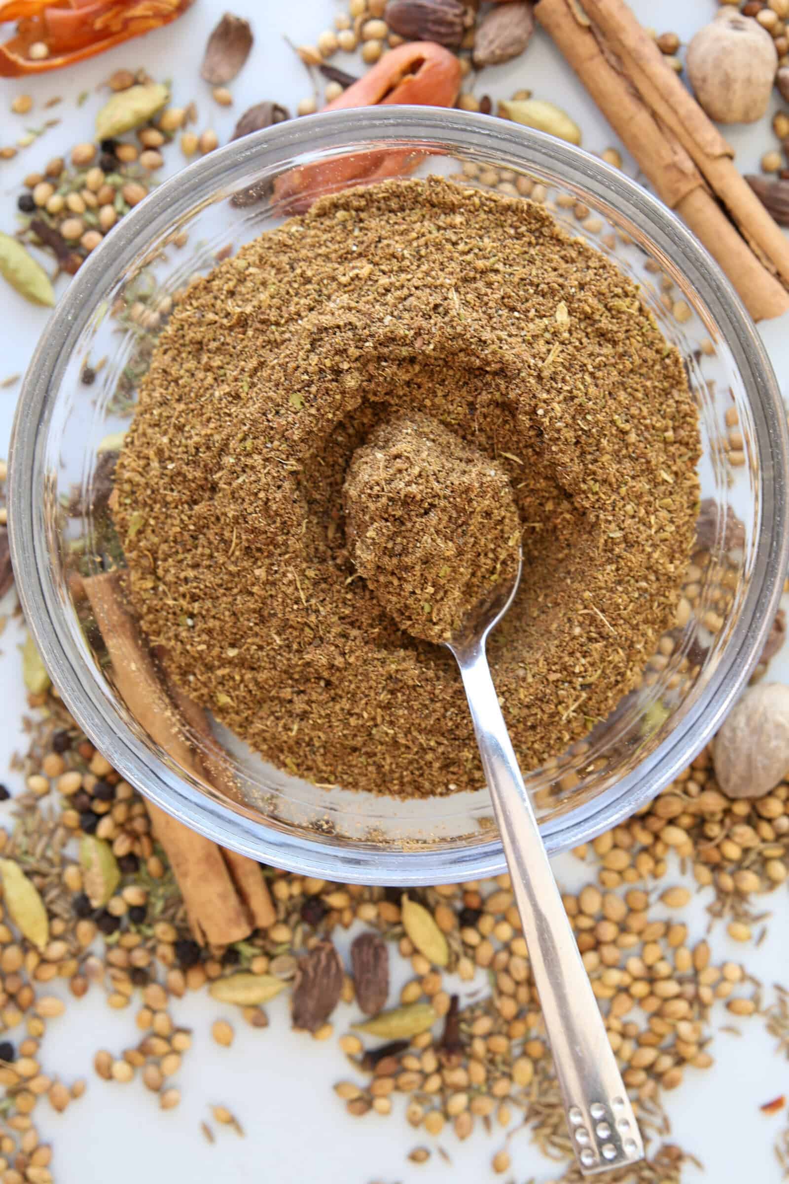 A glass bowl filled with ground spice mix sits on a white surface, surrounded by whole spices like cinnamon sticks, cardamom pods, coriander seeds, and black pepper. A spoon rests in the bowl of spices.