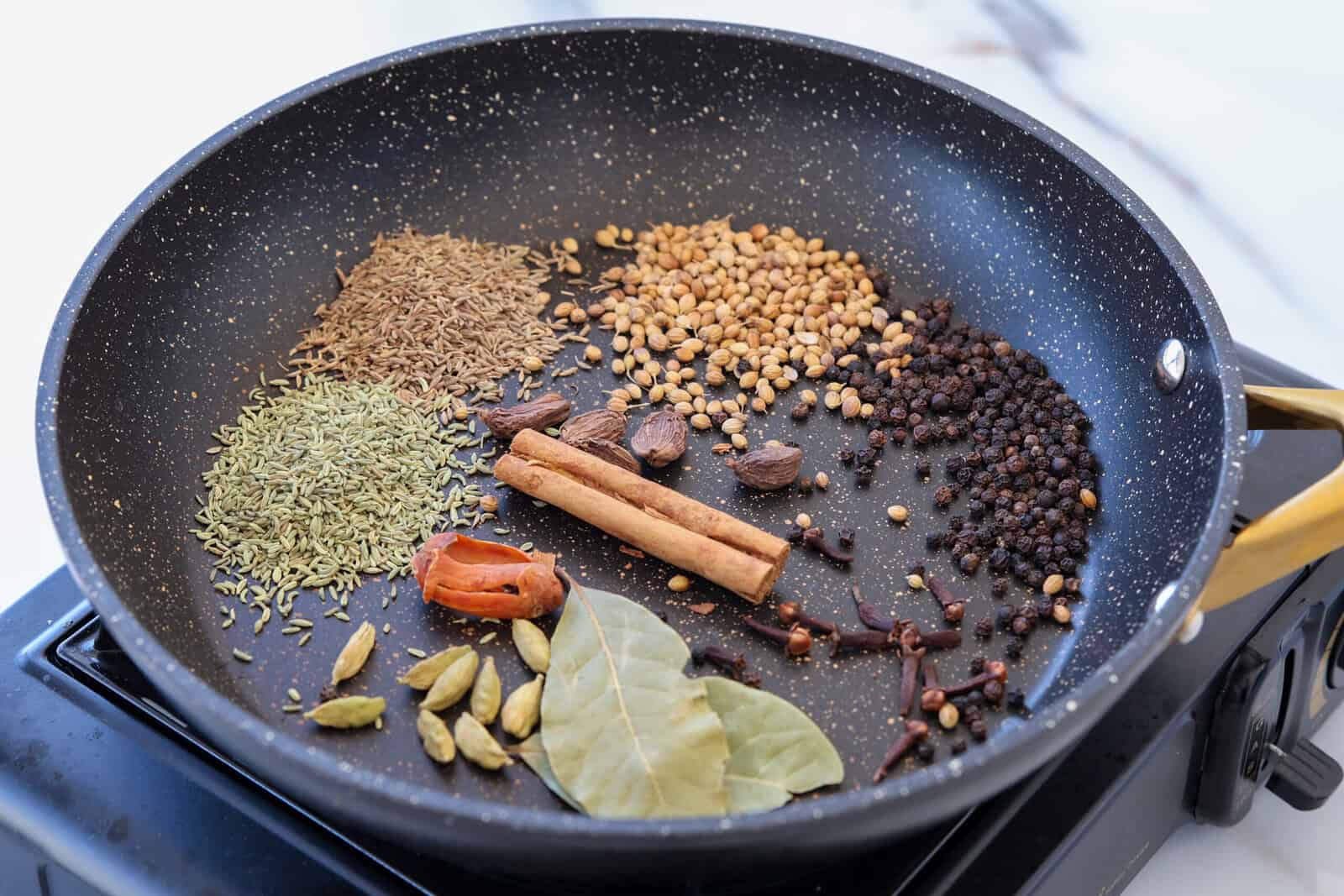 A black frying pan on a stove contains whole spices including cumin, fennel, coriander seeds, black cardamom, cinnamon sticks, black peppercorns, bay leaves, cloves, green cardamom, and a mace blade.