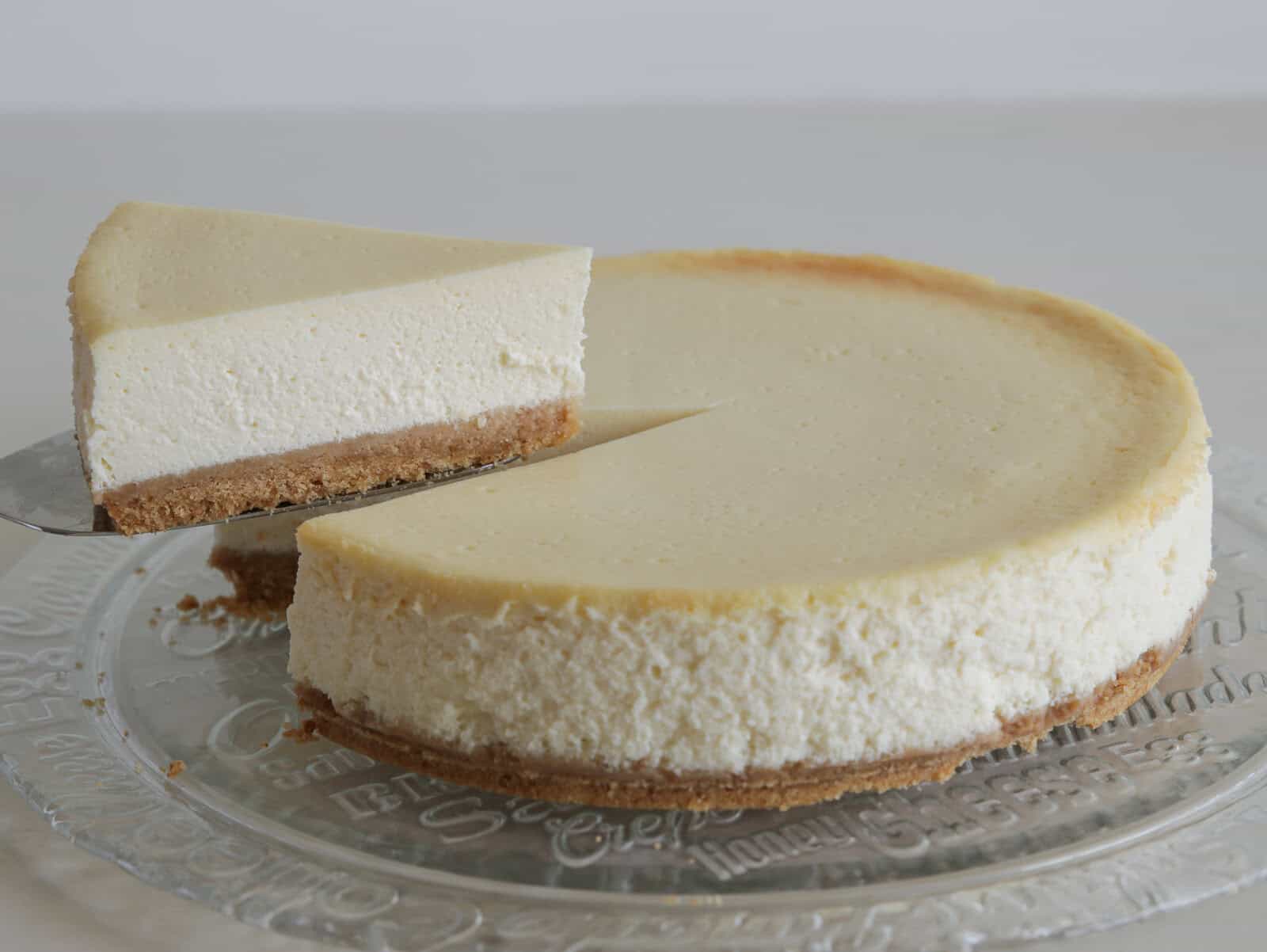 A classic cheesecake with a golden-brown crust on a glass plate, with one slice being lifted out, revealing its creamy, smooth texture and thick graham cracker base.