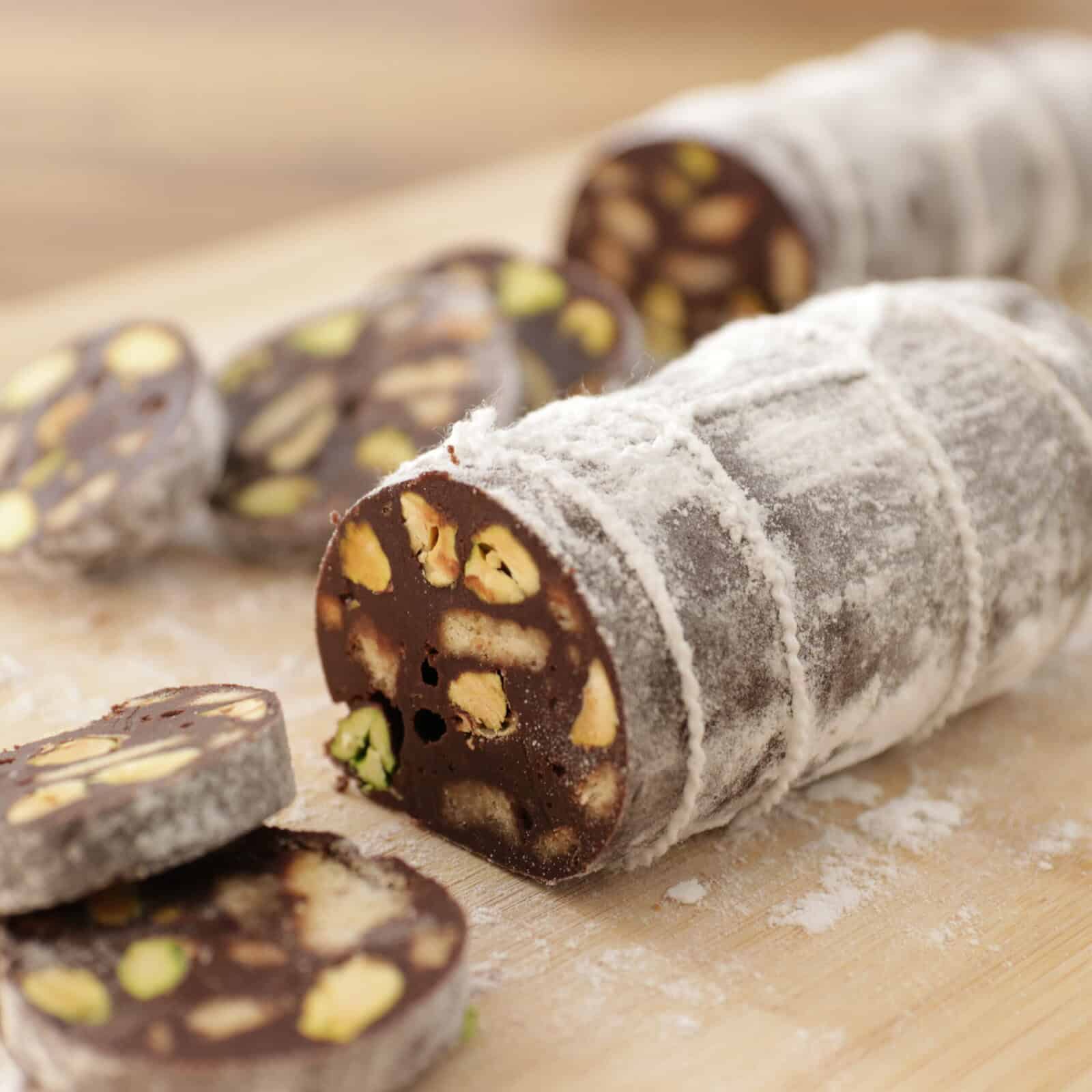 A chocolate salami on a wooden board, dusted with powdered sugar and sliced to reveal pieces of pistachio and other nuts inside.
