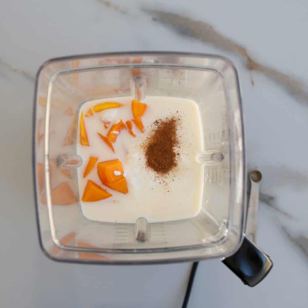 A blender viewed from above contains milk, chunks of orange fruit, and a sprinkle of brown spice on top, set on a white marble countertop.