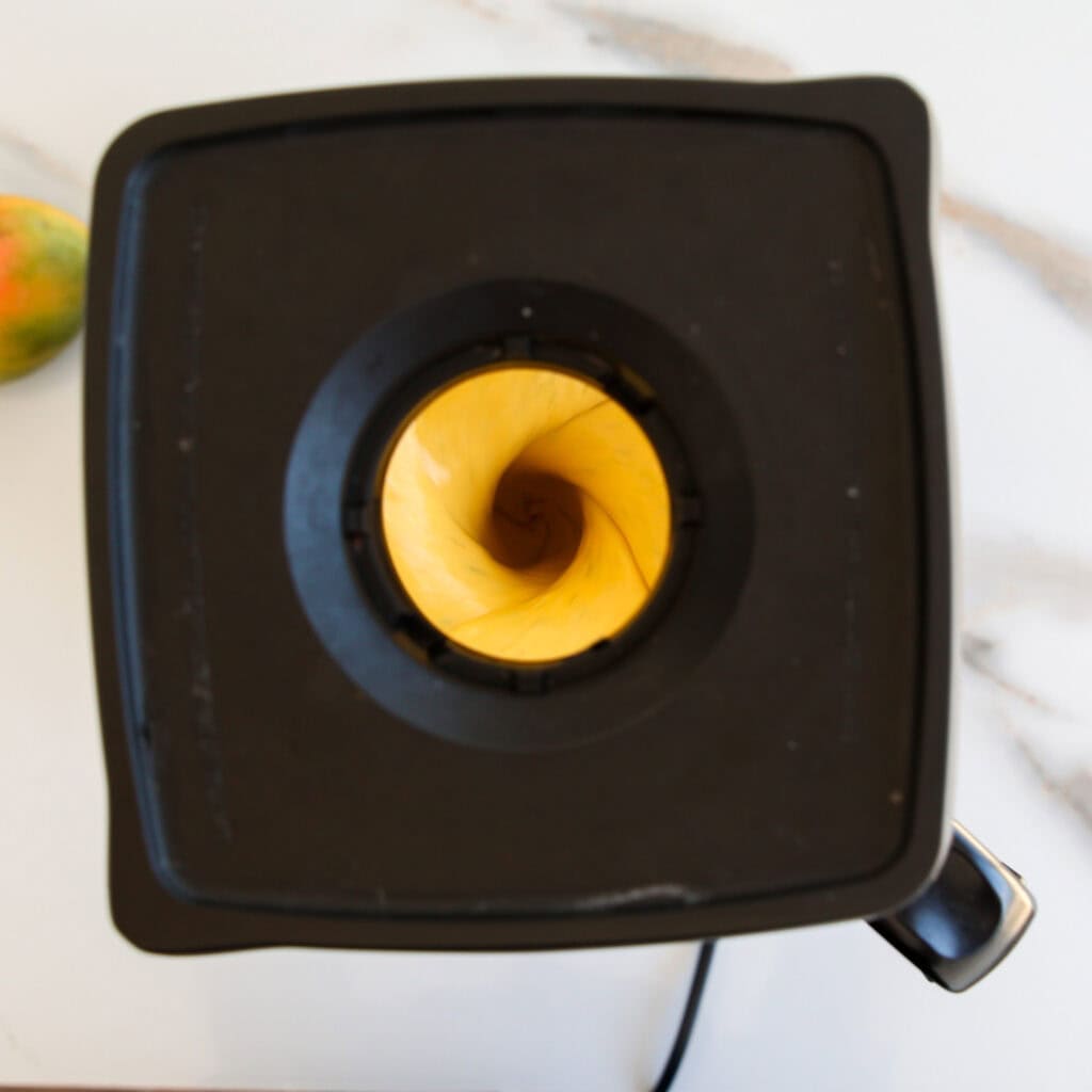 Top view of a blender filled with a creamy yellow mixture, likely a smoothie in the process of blending. The blender sits on a white marble countertop, with part of a mango visible on the left.