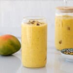 A glass filled with yellow mango lassi, topped with crushed nuts, sits on a white surface. A whole mango, a jar with more lassi, and a small patterned bowl are nearby.