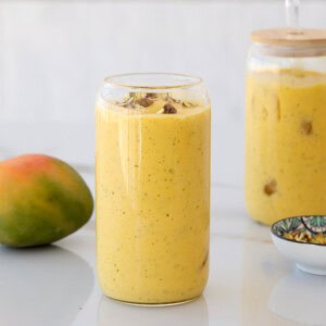 A glass filled with yellow mango lassi, topped with crushed nuts, sits on a white surface. A whole mango, a jar with more lassi, and a small patterned bowl are nearby.