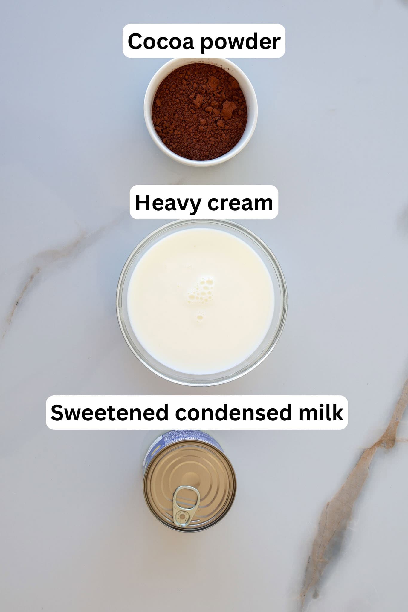 Three ingredients on a marble surface: a small bowl of cocoa powder at the top, a larger bowl of heavy cream in the middle, and an open can of sweetened condensed milk at the bottom.