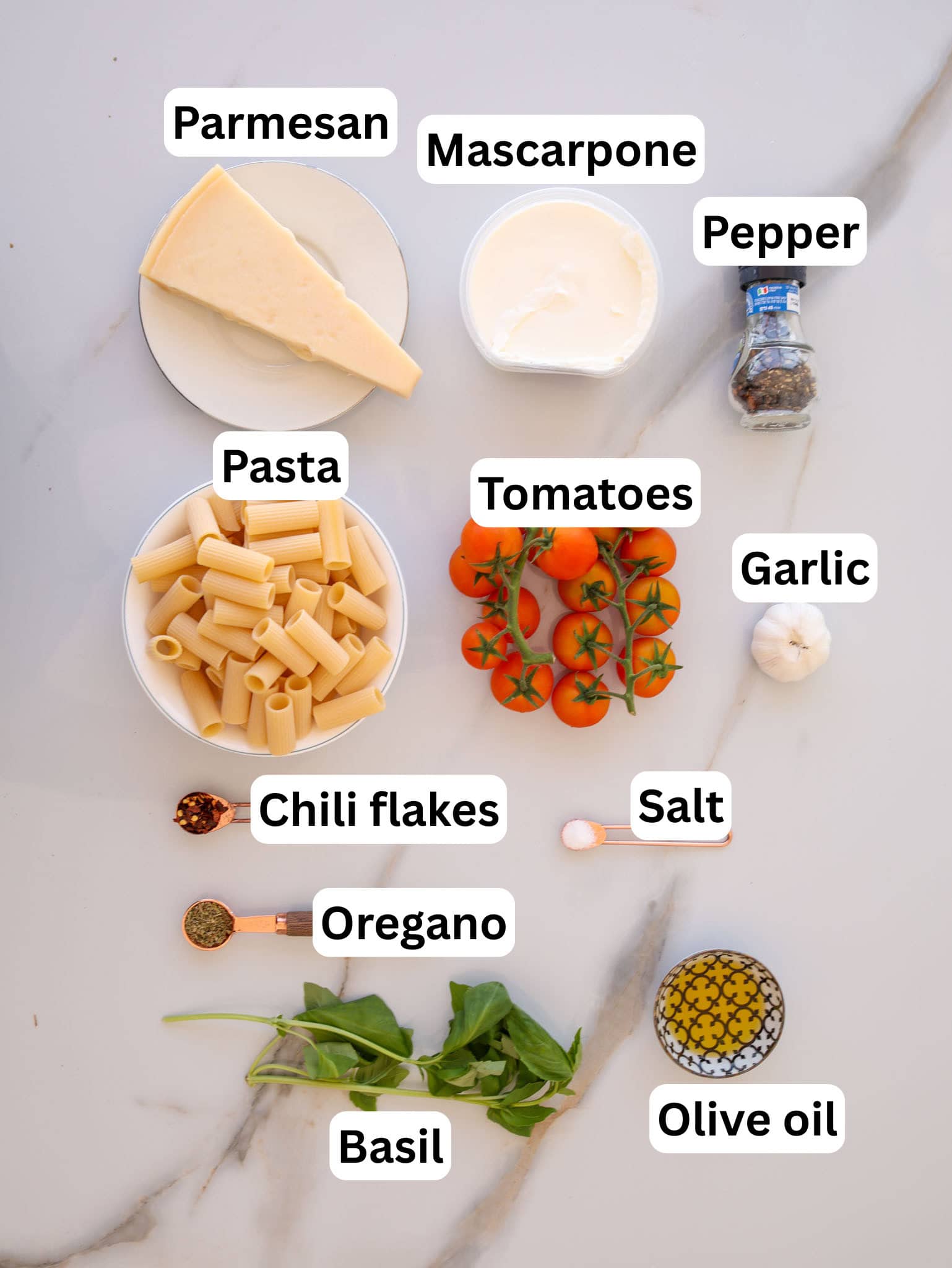 Overhead view of labeled ingredients on a white surface: parmesan, mascarpone, pepper, pasta, tomatoes on the vine, garlic, chili flakes, salt, oregano, olive oil, and fresh basil leaves.