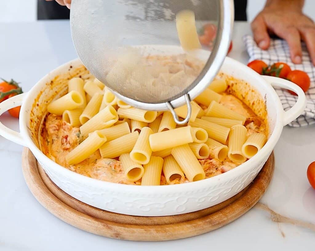 A person pours cooked rigatoni pasta from a strainer into a white baking dish with creamy tomato sauce, set on a wooden board. Fresh tomatoes and a checkered cloth are nearby.