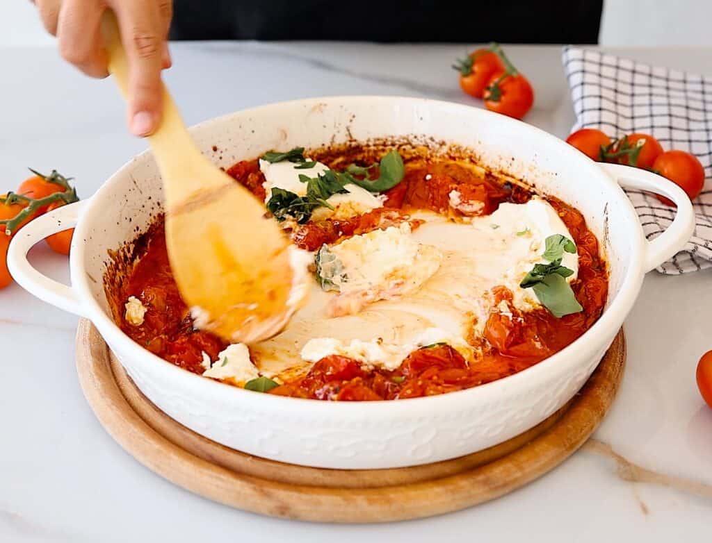 A hand stirs baked feta cheese, tomatoes, and herbs in a white casserole dish with a wooden spoon; cherry tomatoes and a checkered cloth are on the table nearby.