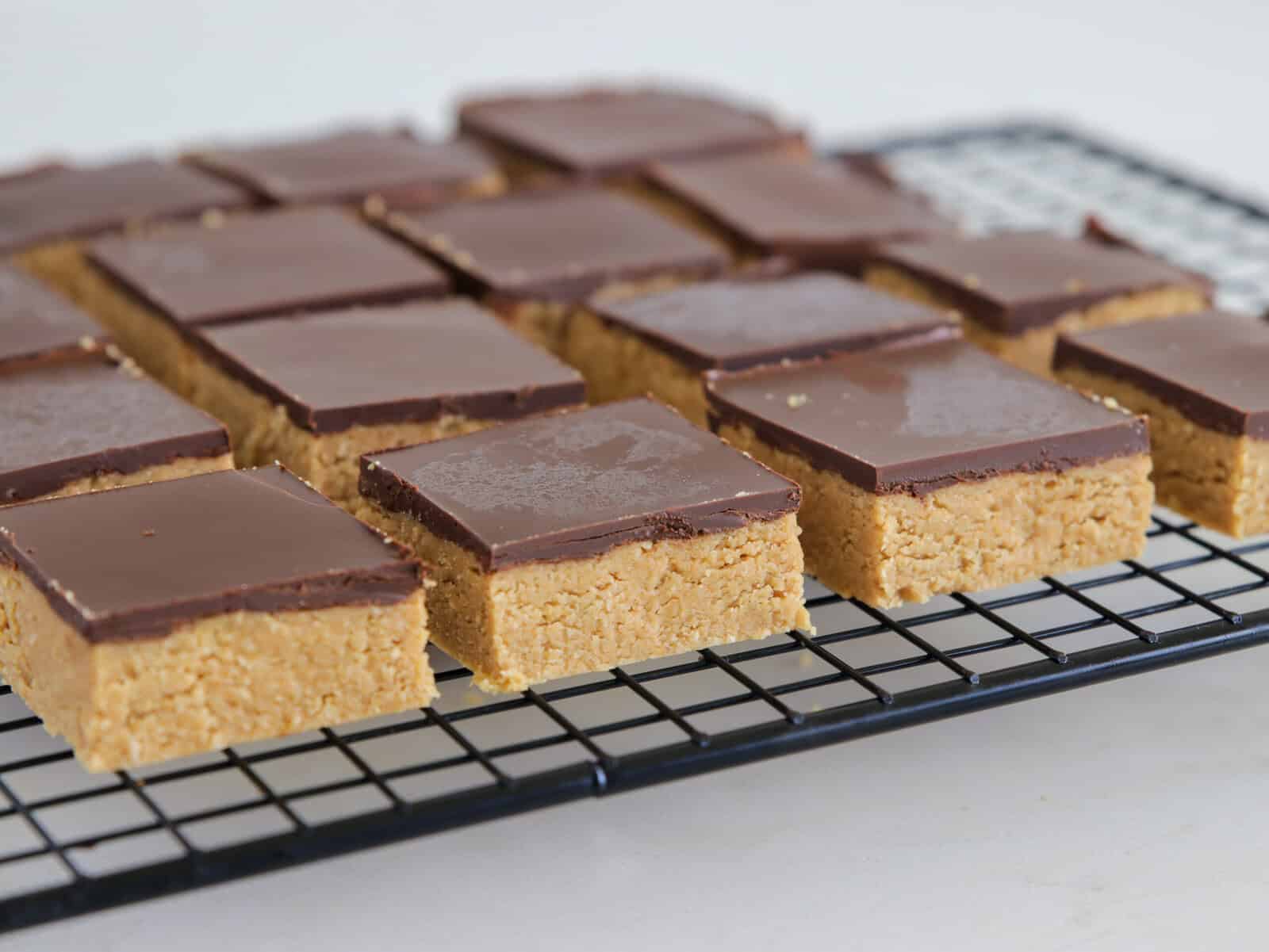 Several square peanut butter bars topped with a layer of chocolate are cooling on a black wire rack. The bars have a smooth, shiny chocolate layer and a dense, light brown base.