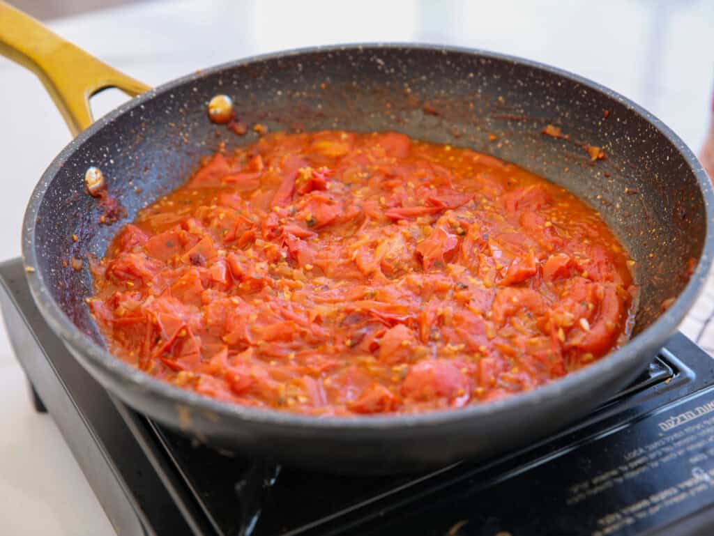 A black frying pan with a yellow handle sits on a stove, filled with simmering tomato sauce made of chopped tomatoes. The sauce appears thick and chunky.