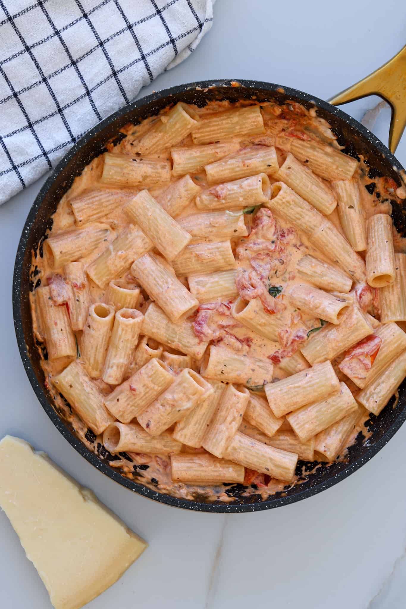 A pan filled with creamy rigatoni pasta in a tomato-mascarpone based sauce, garnished with herbs. A block of cheese and a black-and-white checkered cloth are nearby on a white surface.