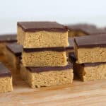 A close-up of peanut butter bars with a chocolate layer on top, stacked and arranged on a wooden surface. The bars have a thick, crumbly peanut butter base and a smooth chocolate topping.