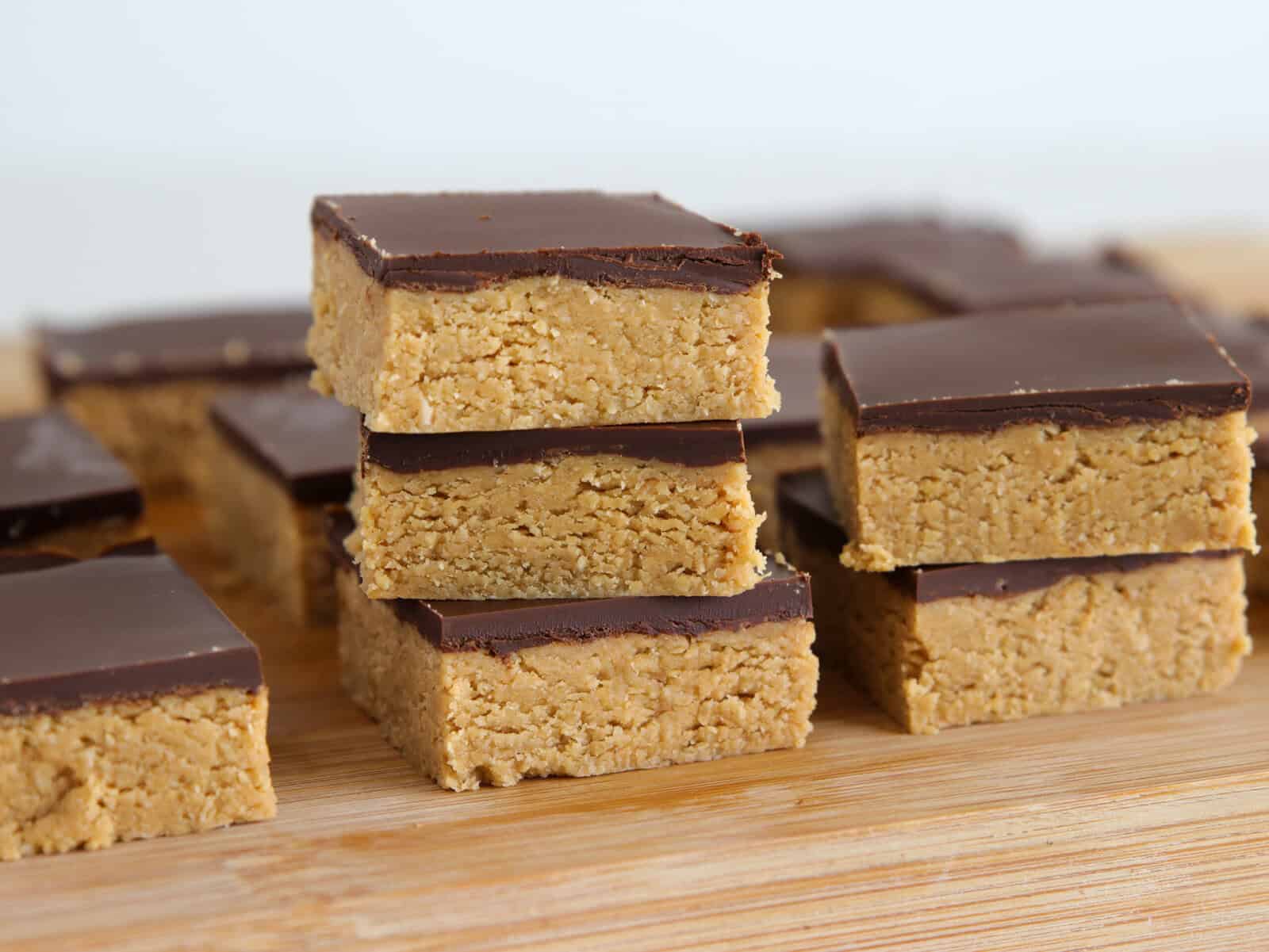 A close-up of peanut butter bars with a chocolate layer on top, stacked and arranged on a wooden surface. The bars have a thick, crumbly peanut butter base and a smooth chocolate topping.