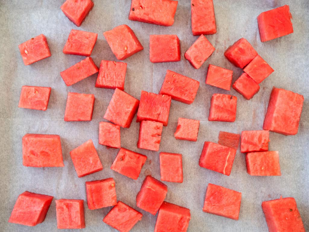 Cubes of fresh, bright red watermelon spread out on a sheet of parchment or wax paper in a scattered arrangement.