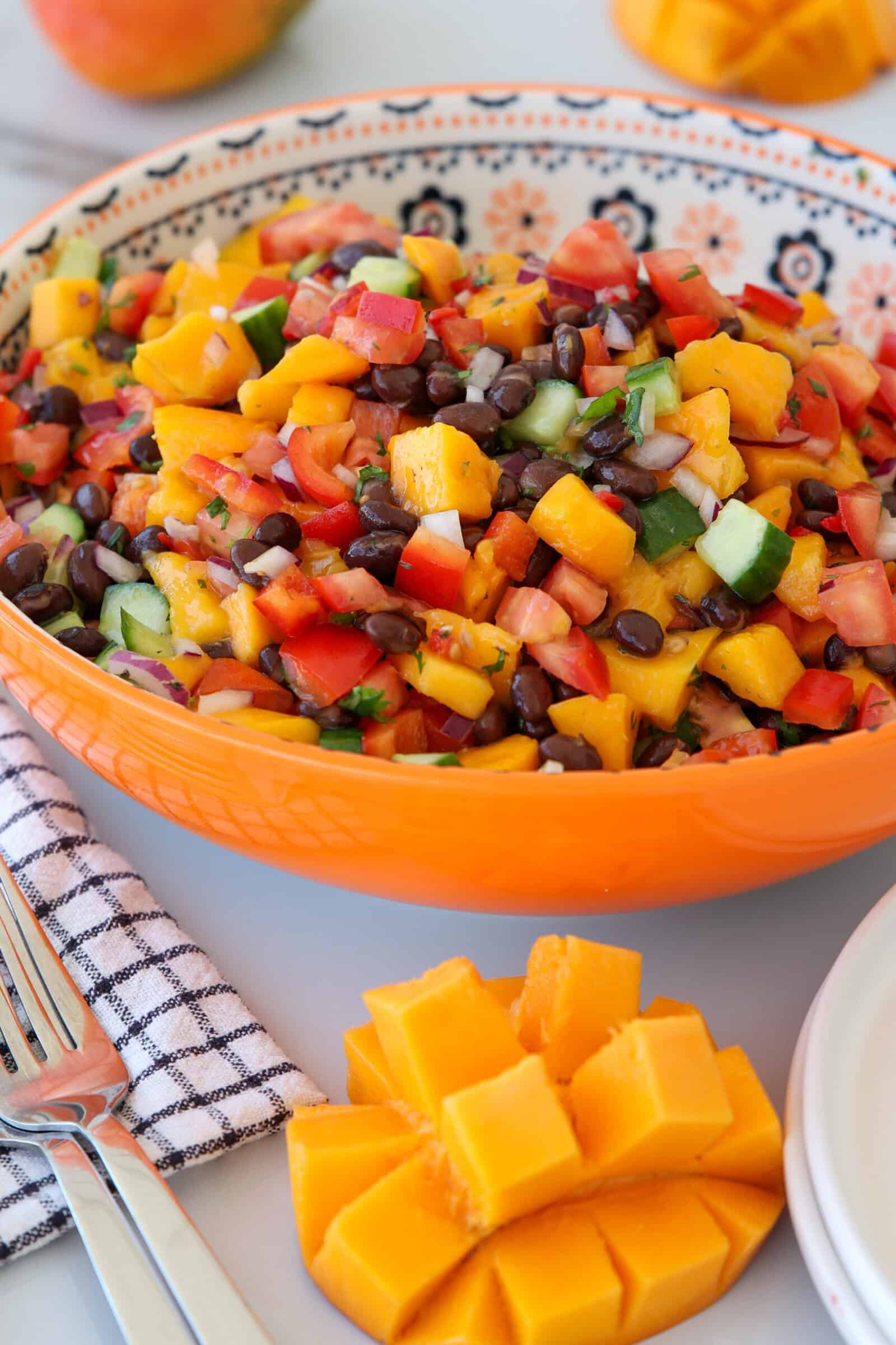 A vibrant bowl of mango black bean salsa with diced mango, black beans, red bell pepper, cucumber, onion, and cilantro. A cubed mango and utensils are beside the bowl on a white surface with a striped napkin.