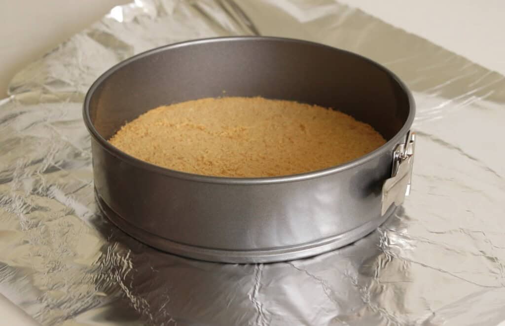 A round springform pan filled with a pressed graham cracker crumb crust sits on top of a sheet of aluminum foil.