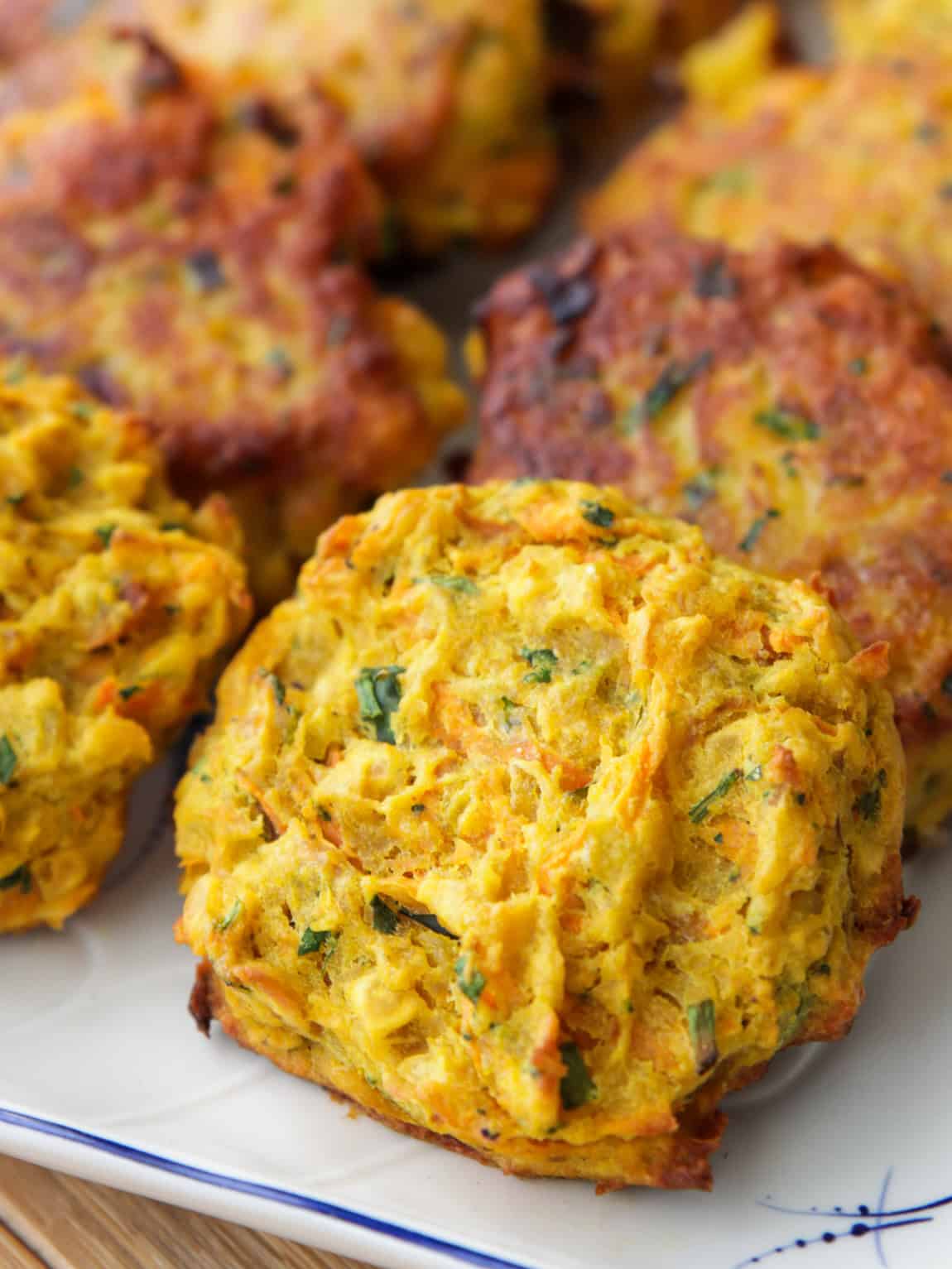 Close-up of several golden-brown lentil patties with visible pieces of green herbs and shredded vegetables, arranged on a white plate.