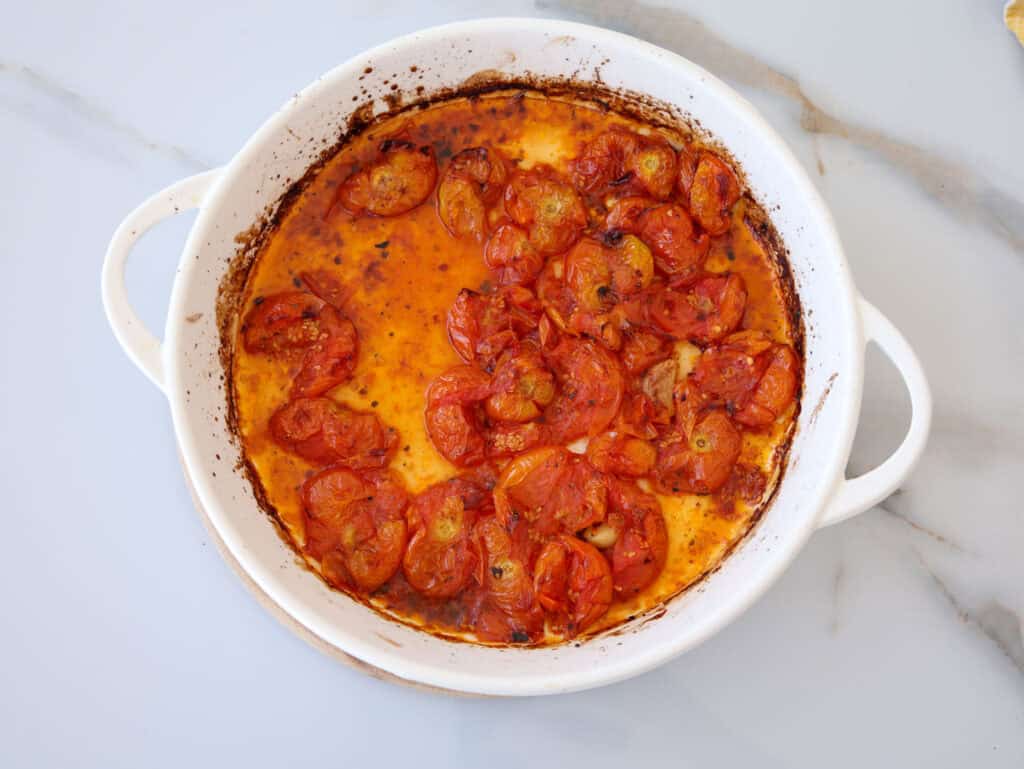 A white round baking dish filled with roasted cherry tomatoes in olive oil, with browned edges and visible seasoning, sits on a light marble surface.