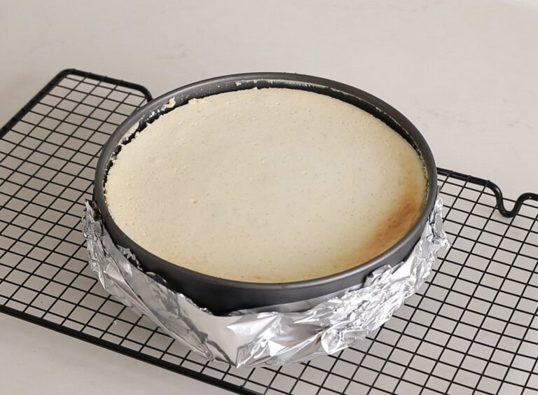 A round cheesecake in a pan, wrapped with aluminum foil, cools on a black wire rack against a light background. The top is smooth with a slight golden tint at the edge.