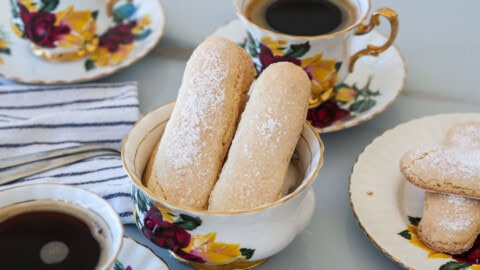 Two sugar-dusted ladyfinger biscuits in a floral teacup, surrounded by matching teacups of black coffee and plates on a table with a striped napkin.