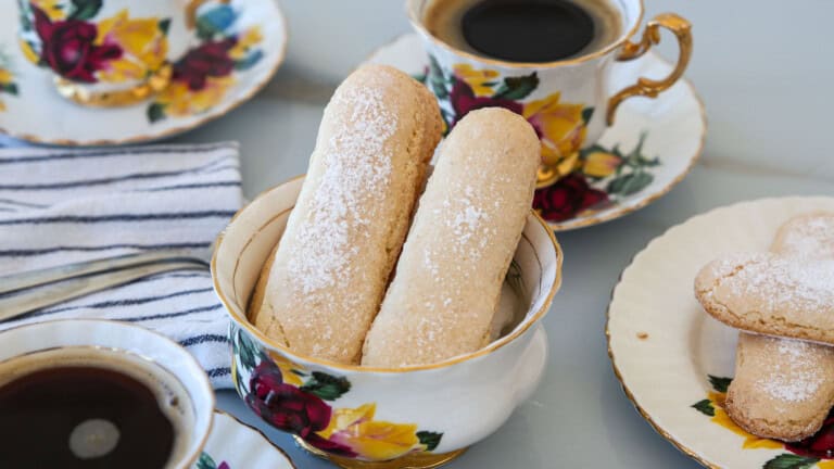 Two sugar-dusted ladyfinger biscuits in a floral teacup, surrounded by matching teacups of black coffee and plates on a table with a striped napkin.