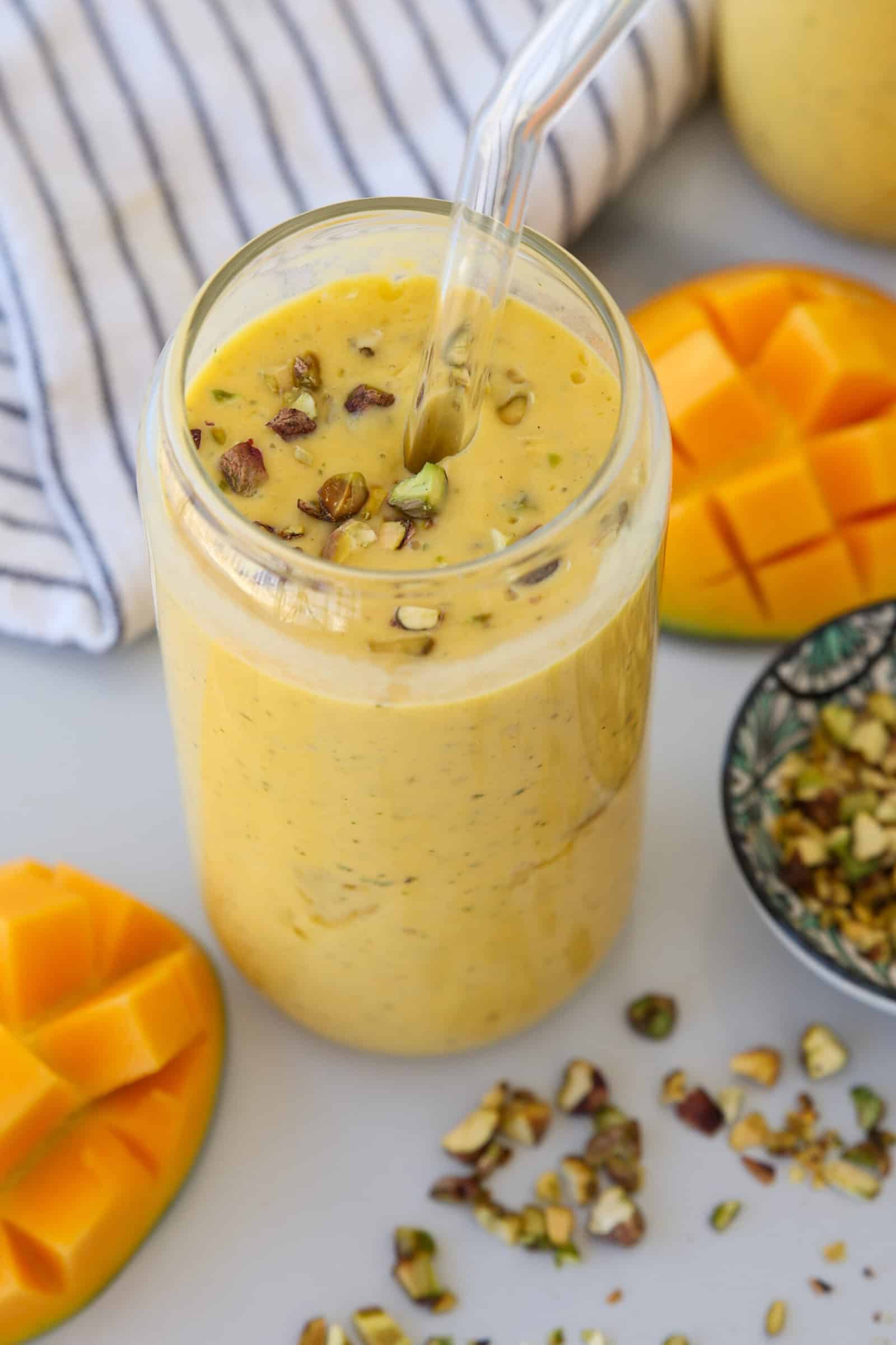 A glass of creamy mango smoothie topped with chopped pistachios, with a clear straw. Sliced mango pieces and a small bowl of chopped pistachios are nearby, and a striped cloth is in the background.