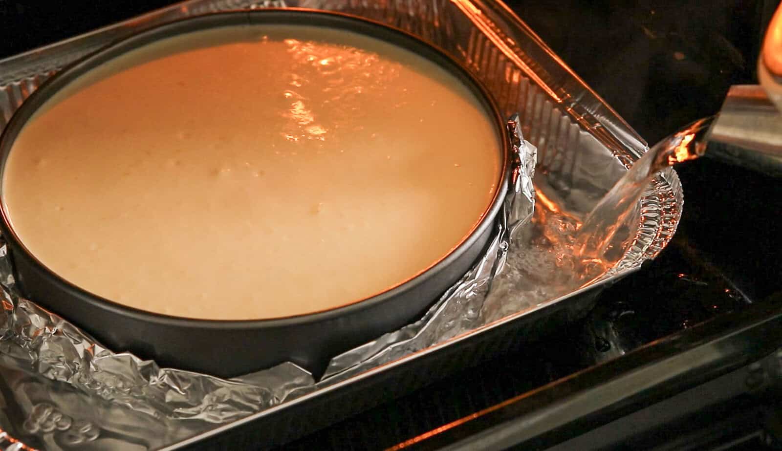 A cheesecake pan wrapped in foil sits in a larger pan in the oven, while water is being poured in to create a water bath for baking.